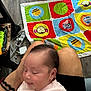 baby, infant, sleeping, person, lap, legs, play_mat, animal_pattern, floor, wood_floor, furniture, bracelet, cables, charger, indoor, clothing, pink_outfit, head, hair, skin