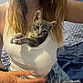 kitten, cat, person, white_tank_top, jeans, bed, bedding, patterned_pillow, hand, long_hair, casual_clothing, indoor, cute, pet, domestic_animal, resting, curious, feline, young_cat, cozy