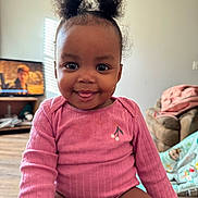Zayanni is registered to the contest to win money with this photo: baby, child, smiling, tongue_out, pink_clothing, onesie, hair_puffs, living_room, television, couch, blanket, floor, wooden_floor, person, cute, portrait, happy, indoor, face, infant