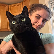 Sissy joined the competition — help win amazing prizes! person, cat, black_cat, indoor, kitchen, wooden_cabinets, holding, pet, wide_eyes, curious, close_up, human, animal, feline, domestic, companion, affection, face, portrait, casual