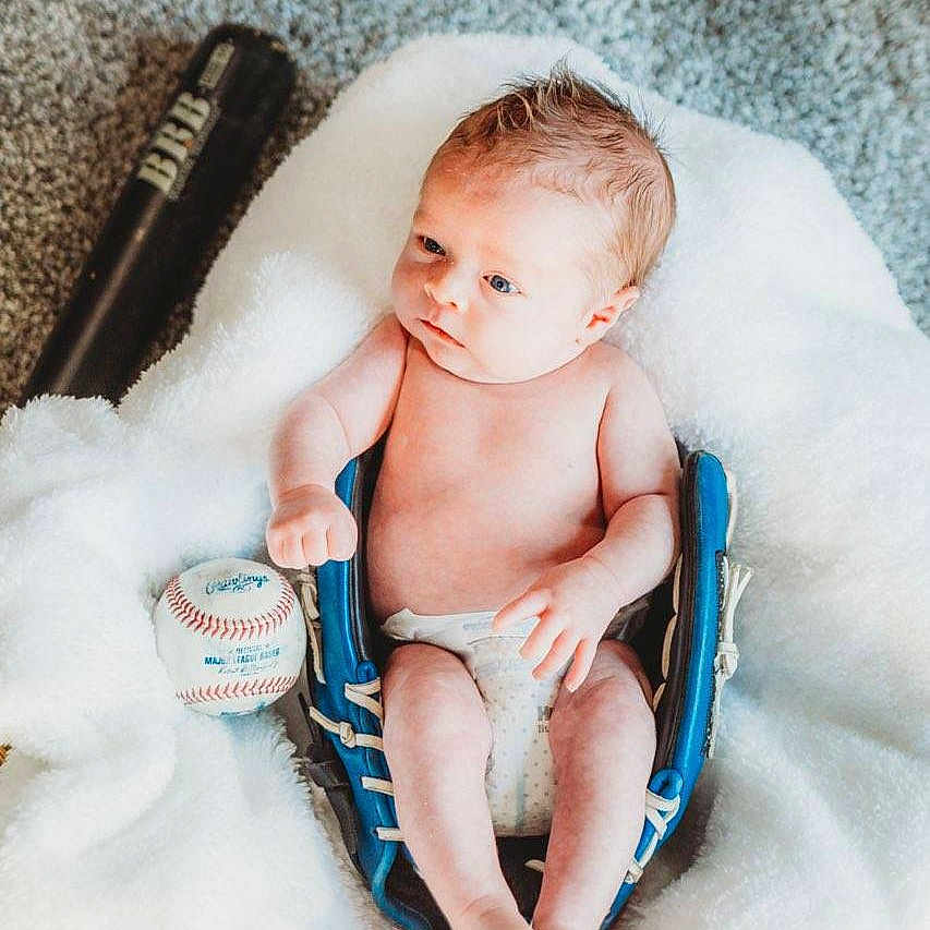 Brixton is registered to the contest to win money with this photo: baby, infant, baseball_glove, baseball, bat, blanket, carpet, diaper, blue, sports_equipment, soft_texture, cute, small, lying_down, indoors, person, newborn, relaxed, fingers, feet