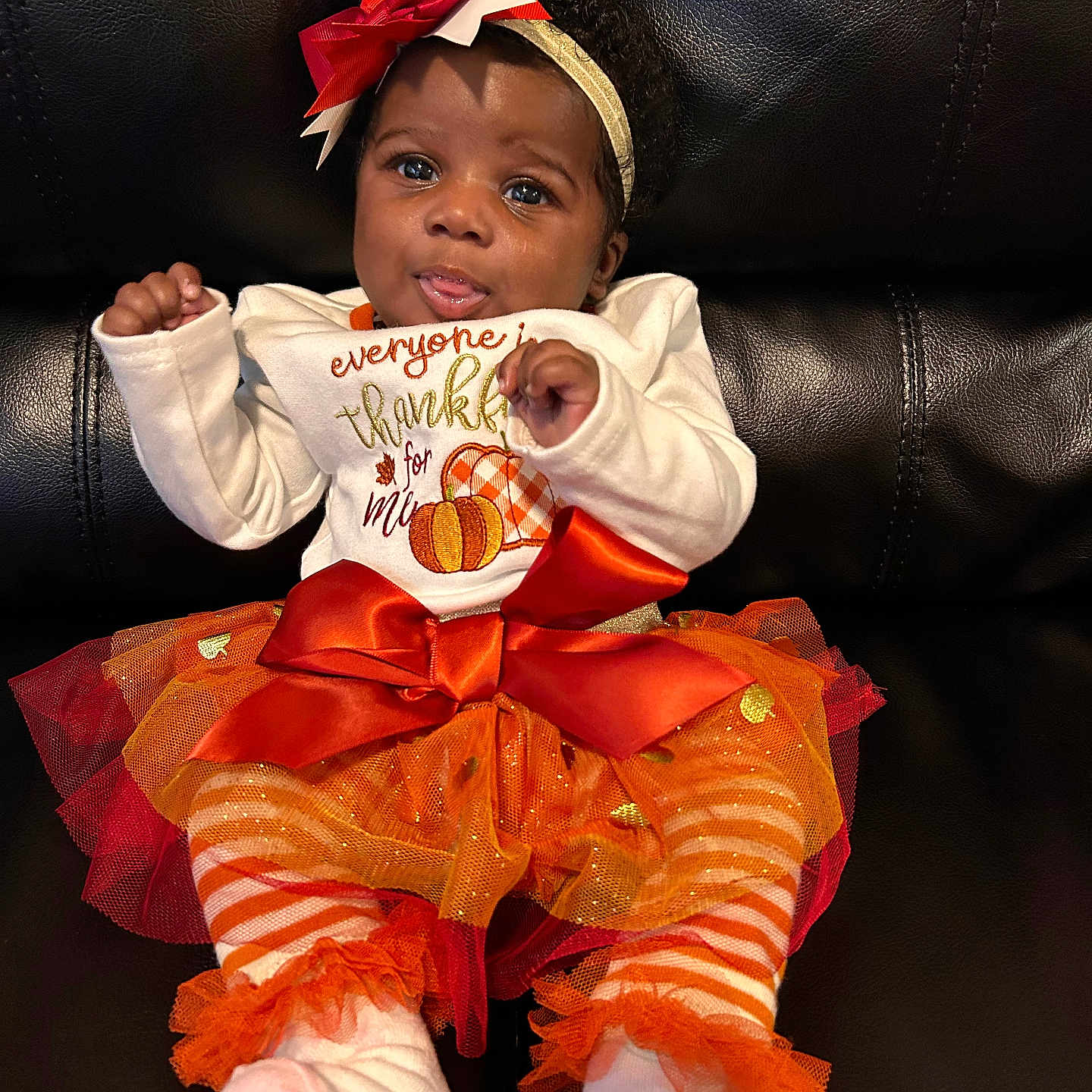 Arnell is registered to the contest to win money with this photo: autumn, baby, black_leather_couch, bow, child, cute, festive, headband, indoor, infant, orange, playful, portrait, pumpkin_design, sitting, smiling, socks, striped_leggings, tutu, white_clothing