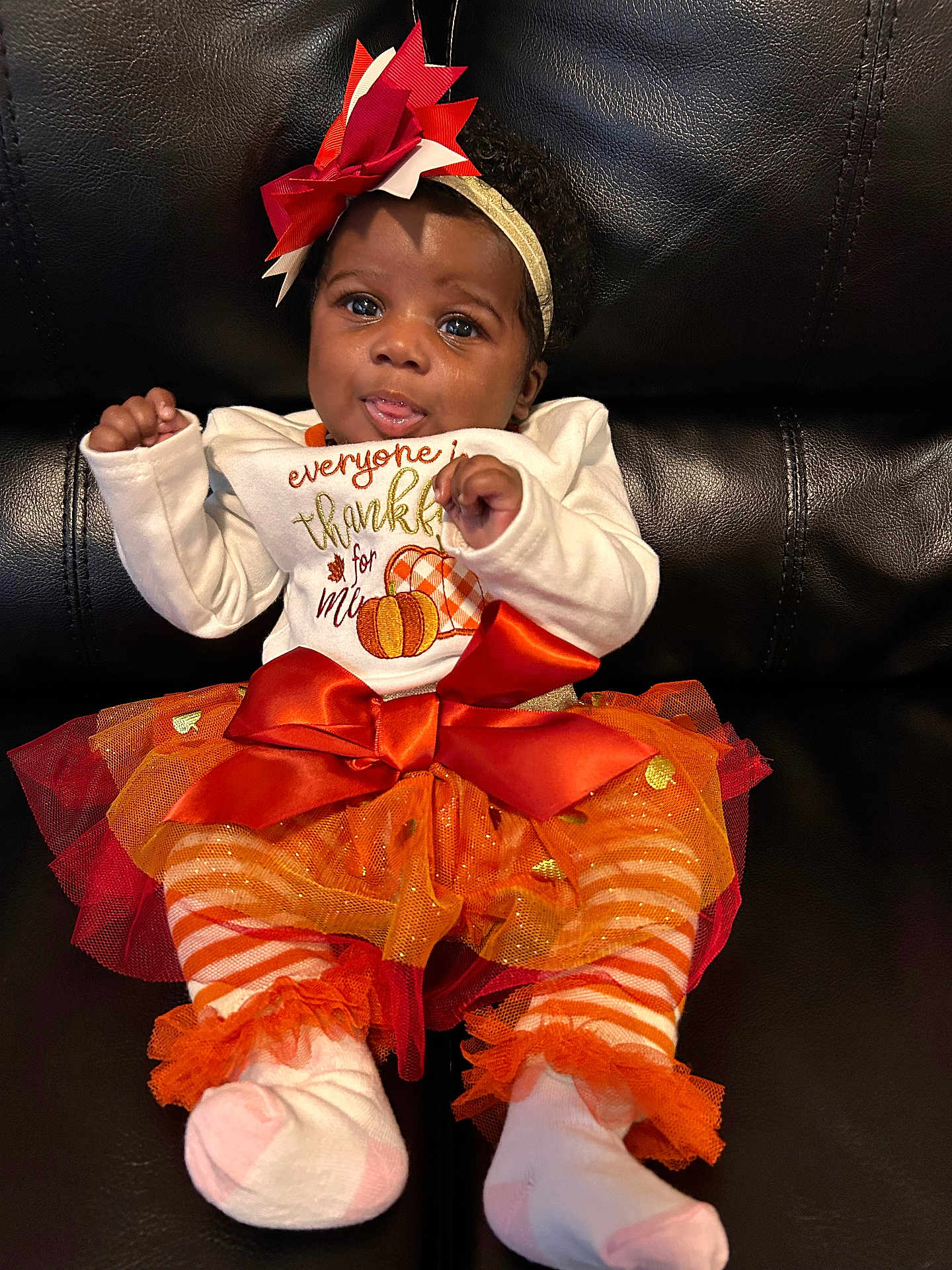 Arnell is registered to the contest to win money with this photo: baby, infant, child, tutu, headband, bow, orange, white_clothing, striped_leggings, socks, black_leather_couch, cute, playful, festive, autumn, pumpkin_design, sitting, portrait, smiling, indoor