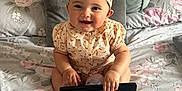 Gabriella joined the competition — help win amazing prizes! baby, child, smiling, headband, curly_hair, remote_control, bed, pillows, floral_bedspread, tufted_headboard, happy, cute, indoors, sitting, clothing, footwear, person, portrait, soft_lighting, decor