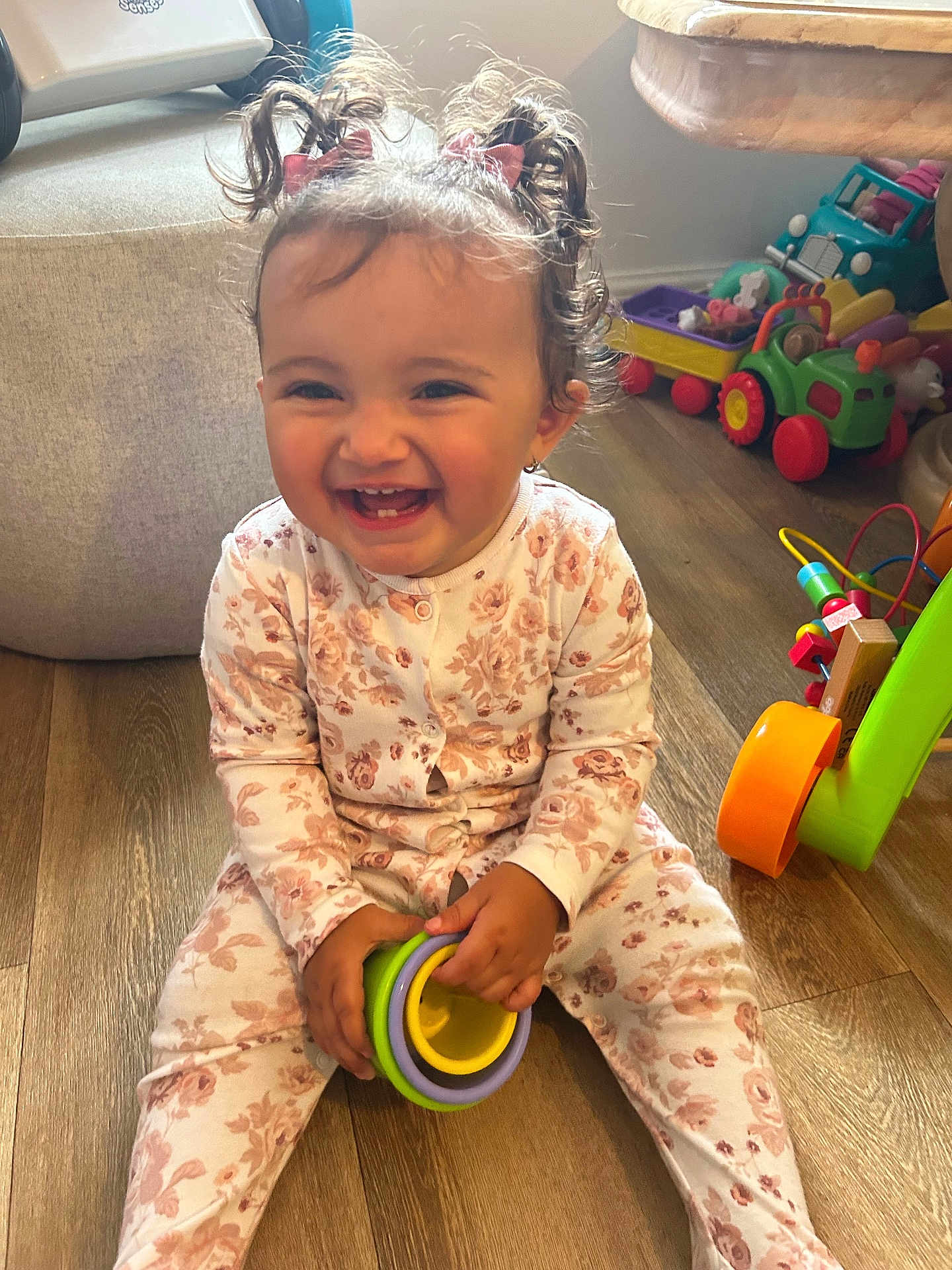 Gabriella is registered to the contest to win money with this photo: baby, bows, child, colorful, curly_hair, cute, floral_pajamas, furniture, happy, indoor, person, pigtails, playroom, playtime, sitting, smiling, stacking_rings, toddler, toys, wooden_floor