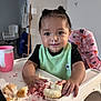 toddler, child, highchair, cupcake, messy, food, frosting, crumbs, pink_cup, hair_buns, green_bib, black_shirt, indoor, table, face, hands, expression, happy, breaking_food, home
