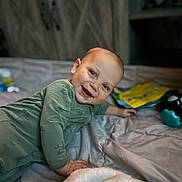 Kouver joined the competition — help win amazing prizes! baby, smile, green_onesie, bed, blanket, toy, indoor, face, child, happy, lying_down, cozy, wooden_background, infant, candid, person, cute, soft_light, playful, portrait