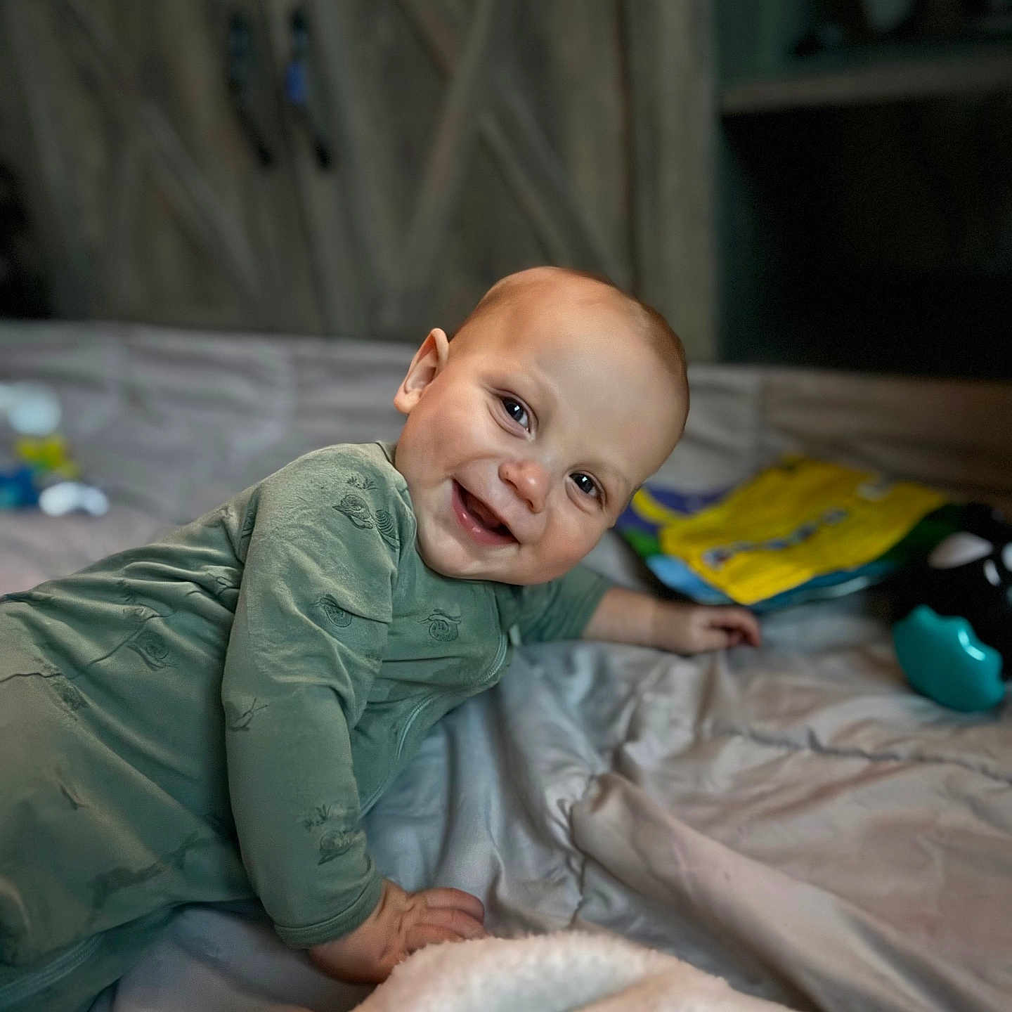 Kouver joined the competition — help win amazing prizes! baby, bed, blanket, candid, child, cozy, cute, face, green_onesie, happy, indoor, infant, lying_down, person, playful, portrait, smile, soft_light, toy, wooden_background