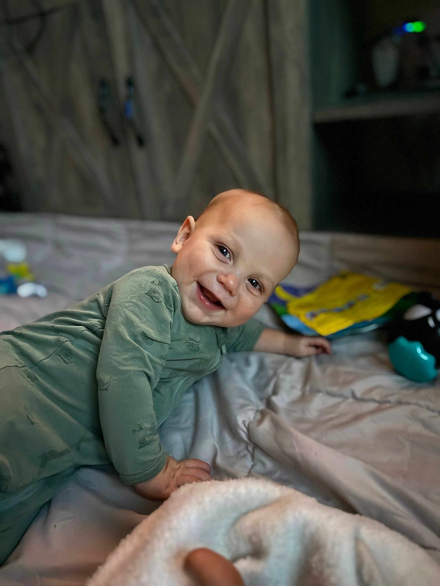 Kouver joined the competition — help win amazing prizes! baby, smile, green_onesie, bed, blanket, toy, indoor, face, child, happy, lying_down, cozy, wooden_background, infant, candid, person, cute, soft_light, playful, portrait