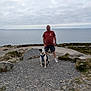 Arley participe au concours pour gagner de l'argent avec cette photo : man, dog, ocean, rocky_path, cloudy_sky, outdoor, casual_clothing, shorts, tshirt, pet, sea, nature, coastline, grass, rocks, leash, standing, sitting, daytime, calm