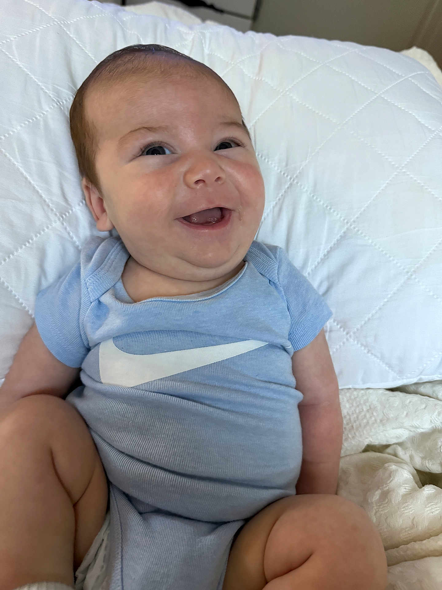 Evan is registered to the contest to win money with this photo: baby, smiling, infant, blue_onesie, white_pillow, quilted, cute, happy, child, person, sitting, indoors, soft_lighting, face, skin, mouth_open, expression, legs, arm, clothing