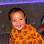 Carmelo is registered to the contest to win money with this photo: baby, child, smiling, orange_clothing, pumpkin_pattern, plaid, bed, couch, happy, indoor, face, head, person, cute, infant, seated, bright_colors, decor, lighting, portrait