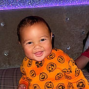 Carmelo is registered to the contest to win money with this photo: baby, child, smiling, orange_clothing, pumpkin_pattern, plaid, bed, couch, happy, indoor, face, head, person, cute, infant, seated, bright_colors, decor, lighting, portrait