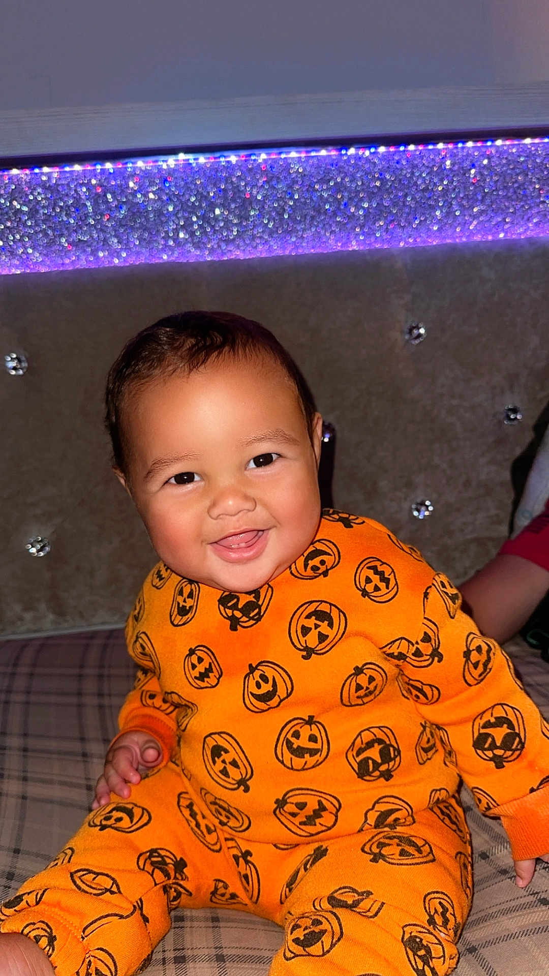 Carmelo is registered to the contest to win money with this photo: baby, child, smiling, orange_clothing, pumpkin_pattern, plaid, bed, couch, happy, indoor, face, head, person, cute, infant, seated, bright_colors, decor, lighting, portrait