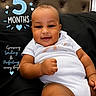 baby, smiling, infant, white_onesie, black_background, headboard, cute, chubby_cheeks, lying_down, indoor, portrait, milestone, text_overlay, celebration, happy, child, young, skin, face, person