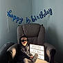 Weston is registered to the contest to win money with this photo: baby, child, sunglasses, hoodie, armchair, sign, celebration, birthday, indoor, feet, smile, light, shadow, banner, text, person, clothing, footwear, happy, milestone