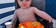 Weston is registered to the contest to win money with this photo: baby, child, orange_shorts, chair, blue_background, striped_background, sun_pattern, summer, outdoor, serious_face, sitting, young_child, bare_chest, light_skin, short_hair, casual, relaxing, daylight, toy, fabric