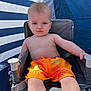 baby, child, orange_shorts, chair, blue_background, striped_background, sun_pattern, summer, outdoor, serious_face, sitting, young_child, bare_chest, light_skin, short_hair, casual, relaxing, daylight, toy, fabric