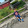 child, boy, smiling, sitting, stone_wall, tshirt, blue_sneakers, outdoor, grass, flowers, barn, trees, fence, sunny, daylight, nature, pathway, casual_clothing, portrait, happy