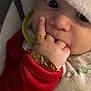 baby, close_up, face, hand, finger, gold_ring, gold_bracelet, hat, red_clothing, bib, sucking_fingers, wide_eyes, soft_texture, indoor, portrait, child, cute, infant, cozy, curious