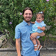 Amelia is registered to the contest to win money with this photo: baby_shoes, beard, blue_shirt, casual_clothing, child, daylight, family, flowers, greenery, happy, holding, man, nature, outdoor, person, plants, portrait, shorts, smiling, toddler