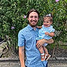 baby_shoes, beard, blue_shirt, casual_clothing, child, daylight, family, flowers, greenery, happy, holding, man, nature, outdoor, person, plants, portrait, shorts, smiling, toddler