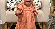 Amelia is registered to the contest to win money with this photo: baby, chair, child, cushion, cute, dress, floral_pattern, furniture, hair_bows, happy, home, indoor, peach_color, portrait, shoes, smiling, socks, standing, toddler, wooden_floor