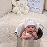 Herick is registered to the contest to win money with this photo: baby, infant, child, portrait, sitting, bucket, metal_container, bonnet, hat, white_clothing, teddy_bears, stuffed_animals, birthday, three_months, sign, couch, pillows, furry_blanket, indoors, smiling