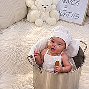 Herick joined the competition — help win amazing prizes! baby, infant, child, portrait, smiling, sitting, bucket, metal_container, teddy_bear, stuffed_animal, nursery, blanket, fluffy_rug, white_clothing, chef_hat, coin, cushions, indoor, milestone_photo, happy