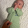 Carson is registered to the contest to win money with this photo: baby, infant, child, lying_down, portrait, face, smiling, hands, fingers, green_outfit, onesie, quilt, bed, bedding, indoors, close_up, adorable, resting, adult_hand, dinosaur_clothing
