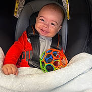 Carson is registered to the contest to win money with this photo: baby, child, infant, smiling, happy, portrait, car_seat, safety_harness, toy, colorful_toy, ball, blanket, red_clothing, gray_clothing, sitting, indoor, vehicle, close_up, face, photography