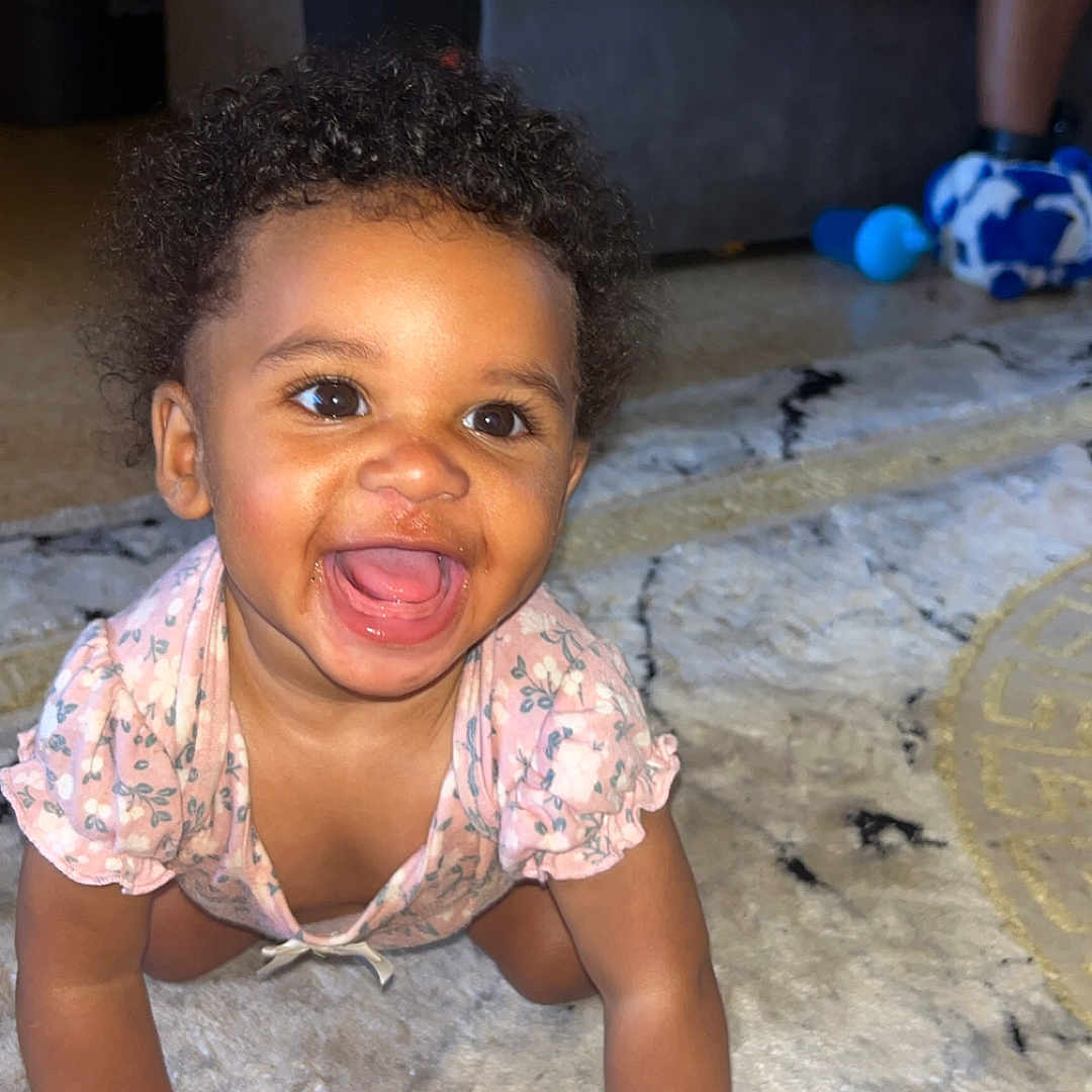Sariyah is registered to the contest to win money with this photo: baby, crawling, smiling, happy, indoor, carpet, child, person, living_room, curly_hair, pink_clothing, cute, toddler, floor, home, young_child, portrait, hands, expression, bright