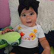 Eliana joined the competition — help win amazing prizes! baby, child, headband, bow, plush_toy, turtle, pillow, bed, pink_wall, black_shorts, white_shirt, floral_pattern, hand, cute, indoor, portrait, smiling, young_child, toy, curious