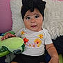 baby, child, headband, bow, plush_toy, turtle, pillow, bed, pink_wall, black_shorts, white_shirt, floral_pattern, hand, cute, indoor, portrait, smiling, young_child, toy, curious