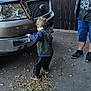 adult, autumn_leaves, casual_clothing, child, concrete, curious, daylight, fence, flag, ford_truck, hoodie, interaction, outdoor, parking, person, sidewalk, sneakers, toddler, tree, vehicle