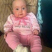 Sophie is registered to the contest to win money with this photo: baby, infant, child, pink_clothing, sitting, couch, pillow, indoor, cute, face, looking, person, sock, long_sleeve, text_on_clothing, soft_fabric, home, comfort, young, portrait