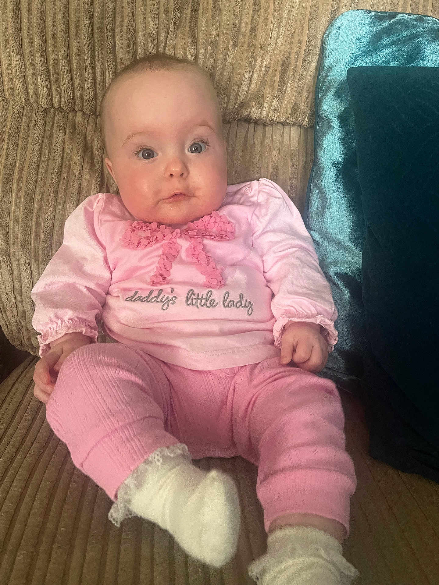 Sophie is registered to the contest to win money with this photo: baby, infant, child, pink_clothing, sitting, couch, pillow, indoor, cute, face, looking, person, sock, long_sleeve, text_on_clothing, soft_fabric, home, comfort, young, portrait