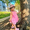 apartment_building, child, curious, daylight, footwear, grass, hat, nature, outdoor, person, pink_crocs, pink_dress, playful, sidewalk, standing, straw_hat, sunny, toddler, tree, young_child