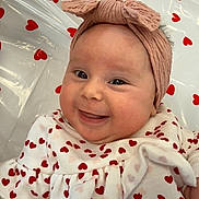 Leighton is registered to the contest to win money with this photo: baby, smile, headband, pink, heart_pattern, clothing, infant, happy, cute, face, cheeks, eyes, lying_down, fabric, pattern, child, portrait, indoor, skin, apparel