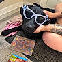 accessory, arm, black_dog, close_up, dog, floor, hand, indoor, mat, pet, plastic_box, playful, relaxed, resting, rubber_bands, snack_pack, stitch, sunglasses, tattoo, wood_floor