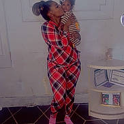 Trevon is registered to the contest to win money with this photo: baby, infant, child, caregiver, woman, kiss, affection, family, portrait, indoor, pajamas, plaid, striped_clothing, heart_stickers, smiling, standing, holding_baby, carpet, side_table, home
