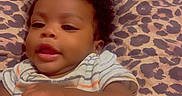 Trevon is registered to the contest to win money with this photo: baby, infant, child, portrait, lying_down, bed, bedspread, leopard_print, curly_hair, dark_skin, striped_onesie, onesie, face, eyes, mouth, hands, indoor, bedroom, soft_lighting, relaxed