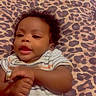 baby, infant, child, portrait, lying_down, bed, bedspread, leopard_print, curly_hair, dark_skin, striped_onesie, onesie, face, eyes, mouth, hands, indoor, bedroom, soft_lighting, relaxed