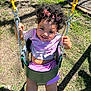 Brixton is registered to the contest to win money with this photo: toddler, child, swing, playground, smiling, curly_hair, pink_bows, outdoor, sunlight, grass, dirt, yellow_chains, happy, casual_clothing, purple_shorts, white_shoes, holding_chains, play, fun, portrait