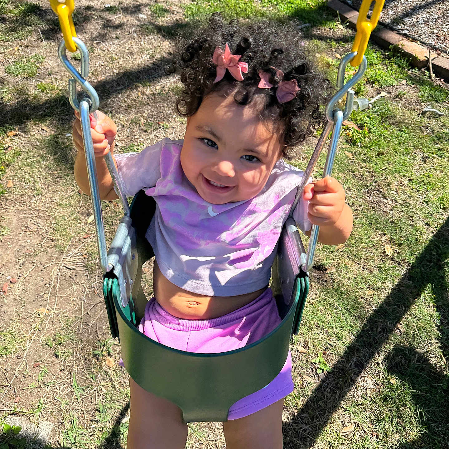 Brixton is registered to the contest to win money with this photo: casual_clothing, child, curly_hair, dirt, fun, grass, happy, holding_chains, outdoor, pink_bows, play, playground, portrait, purple_shorts, smiling, sunlight, swing, toddler, white_shoes, yellow_chains