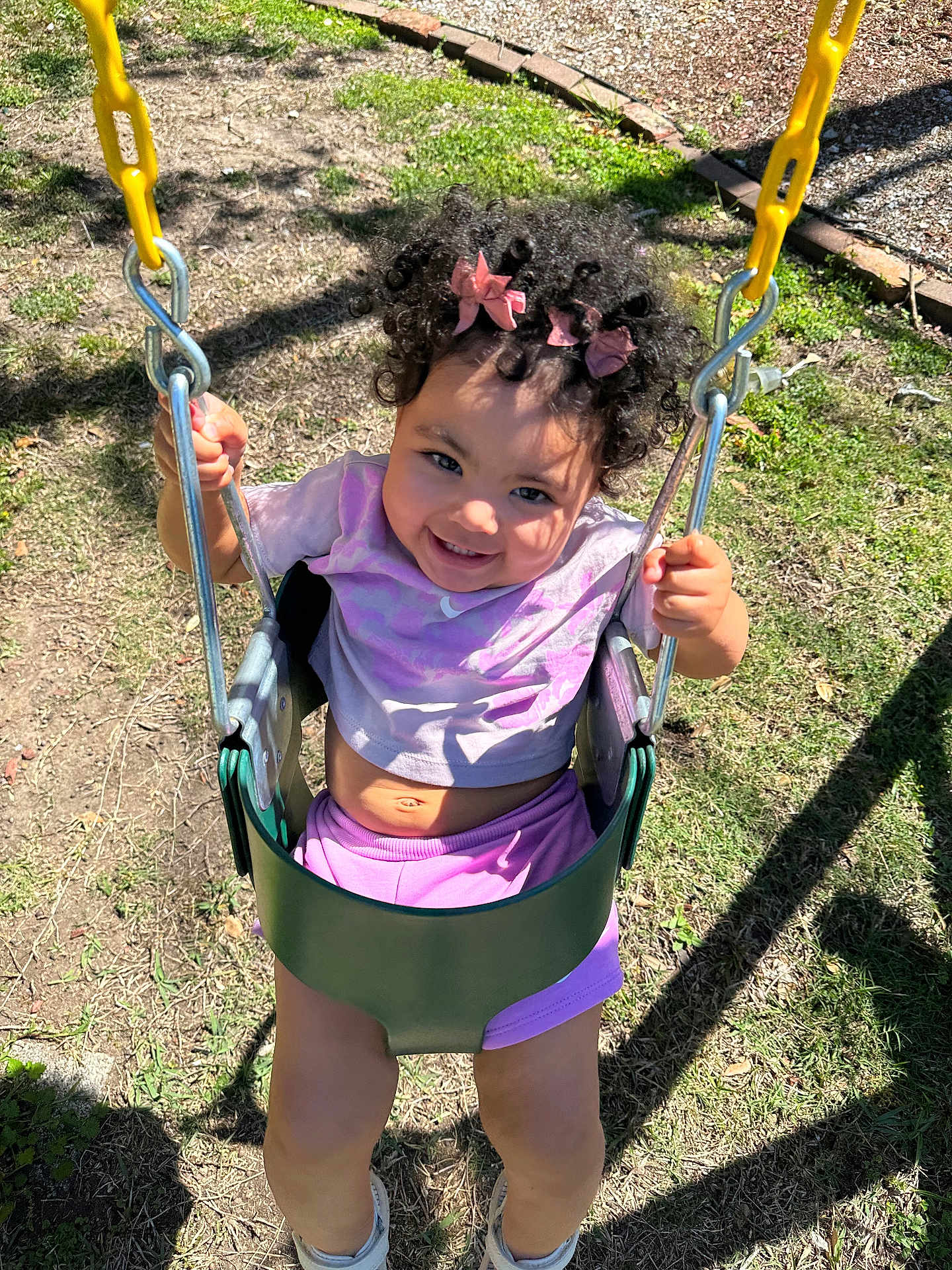 Brixton is registered to the contest to win money with this photo: toddler, child, swing, playground, smiling, curly_hair, pink_bows, outdoor, sunlight, grass, dirt, yellow_chains, happy, casual_clothing, purple_shorts, white_shoes, holding_chains, play, fun, portrait