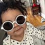 Brixton is registered to the contest to win money with this photo: toddler, child, sunglasses, curly_hair, polka_dot, indoor, flooring, shelf, wooden_floor, clothing, cute, smile, face, person, baby, home, toy, blanket, furniture, portrait
