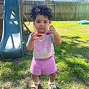 Brixton joined the competition — help win amazing prizes! toddler, child, curly_hair, hair_bow, sunglasses, pink_shorts, pink_shirt, sandals, grass, backyard, slide, fence, daylight, outdoor, person, standing, playground, summer_clothing, shoes, cute