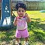 toddler, child, curly_hair, hair_bow, sunglasses, pink_shorts, pink_shirt, sandals, grass, backyard, slide, fence, daylight, outdoor, person, standing, playground, summer_clothing, shoes, cute