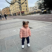 Layan a rejoint le concours — aidez-le/la à gagner de superbes lots ! toddler, child, pink_jacket, white_sneakers, sidewalk, urban, buildings, street, holiday_decorations, pigtails, smiling, outdoor, cold_weather, pavement, people_in_background, cityscape, casual_clothing, daytime, happy, standing